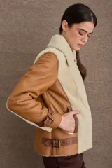 Chaqueta tipo gamulán de cuero color camel con interior y solapas de piel sintética en color beige. Presenta cierre frontal cruzado, correas ajustables con hebillas en los puños y en la cintura, y diseño de cuello solapa.