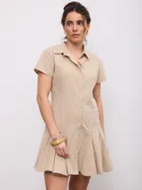 Vestido corto beige de corte camisero, con cuello solapa, mangas corta y cierre frontal con botones a presión. Cuenta con falda evasé con pliegues.