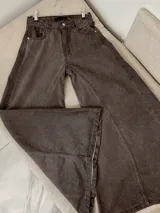 Jean de tiro alto con corte wide leg, confeccionado en denim con efecto lavado en tono marrón oscuro. Presenta costuras frontales verticales que recorren el largo de las piernas, cierre de botón y cremallera, y bolsillos laterales.
