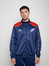 Conjunto deportivo Umbro del Club Nacional de Football, compuesto por una campera con cierre frontal y pantalón a juego. La campera es de color azul con detalles en rojo en los hombros y líneas blancas, presentando el logo de la marca y el escudo del club en el pecho. El conjunto está confeccionado en tejido sintético con acabado satinado.