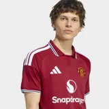Camiseta de fútbol Adidas del Manchester United, modelo Home 26/27. Diseño en color rojo con cuello polo y puños a rayas en blanco y negro, con detalles de las tres tiras de Adidas en los hombros y el logo de Snapdragon en el frente.