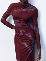 Vestido midi de microtul con diseño drapeado lateral, cuello alto y mangas largas. Presenta un estampado tipo tie-dye en tonos bordó y violeta.