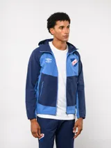 Campera deportiva Umbro del Club Nacional de Football, modelo Summit. Presenta un diseño con bloques de color en azul, rojo y blanco, con capucha, cierre frontal y bolsillos laterales con cierre. Incluye el logo de la marca y el escudo del club en el pecho.