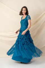 Vestido largo de tul color petróleo, con escote redondo y mangas cortas. Presenta un cuerpo bordado con lentejuelas al tono y una falda con movimiento decorada con apliques de hojas de tela.
