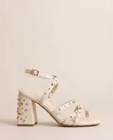 Sandalias de taco ancho de 7 cm, color crema, con tiras cruzadas y cierre de hebilla al tobillo. El diseño presenta múltiples tachas decorativas metálicas en tonos dorados y plateados distribuidas a lo largo de las tiras y en todo el taco.