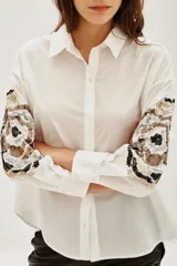 Camisa blanca de algodón con mangas largas, destacada por bordados decorativos en color negro y plateado en las mangas.