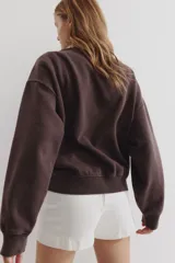 Buzo de tejido de punto color marrón oscuro con efecto lavado, cuello redondo y hombros caídos. Presenta un estampado frontal con el texto "MILANO" en tipografía universitaria, un escudo con la inscripción "193" y la palabra "ACADEMIA" debajo.