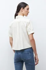 Camisa de algodón color beige claro, con cuello camisero, botones frontales y mangas cortas abullonadas con elástico en el puño.