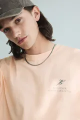 Remera color durazno de corte holgado con cuello redondo y estampado pequeño en el pecho con un tenista y texto.