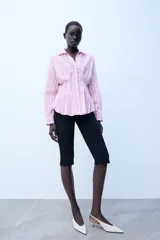 Camisa de popelina con diseño a rayas verticales rosadas y blancas, cuello solapa con escote en V, manga larga y detalle de pliegues marcados en la cintura. Cierre frontal con botones ocultos.