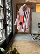 Campera con capucha confeccionada en tejido tipo corderito color rosa, con diseño de collage geométrico en el frente que incluye bloques de color en naranja, negro y blanco. Presenta mangas largas de corte holgado y diseño artesanal único.