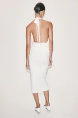 Vestido midi blanco de cuello halter, confeccionado con mezcla de poliamida. Presenta detalle de drapeado lateral con un broche metálico dorado en forma de caracola y cierre en la espalda ajustable con lazadas.