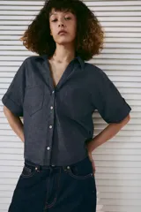 Camisa crop efecto denim color negro, con cuello solapa, escote en V, mangas cortas con trabilla y bolsillos frontales de parche. Cierre frontal con botones.