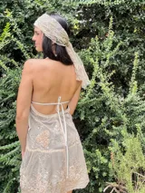 Vestido corto negro con escote en V profundo, estilo halter con tiras para atar al cuello. Presenta un diseño ajustado en el busto con tiras ajustables bajo el mismo que se cruzan y anudan en la espalda, y falda con volado en el ruedo con detalle de encaje.