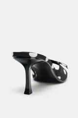 Sandalia tipo mule negra con estampado de lunares blancos, efecto satinado, punta fina, tacón fino y tira delantera con lazo decorativo.