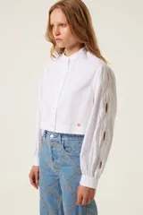 Camisa blanca corta de corte recto con cuello mao, mangas anchas con puños ajustados y aberturas trenzadas en las mangas. Incluye medalla de bronce con logo "TRS".