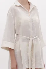 Camisa larga fluida de color crema con cierre frontal de botones, mangas tres cuartos anchas y cinturón ajustable para anudar en la cintura.