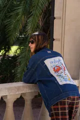 Campera de jean azul oscuro con corte oversize. Presenta un parche rectangular de tela blanca en la espalda con un estampado artístico de temática marina que incluye cangrejos, limones y la frase 'vie légère' sobre un fondo de cuadrícula azul.