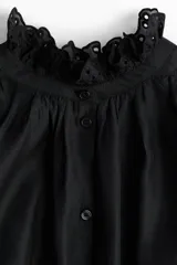 Blusa negra de tejido transparente con cuello alto y puños con volado y detalles de broderie. Presenta frunces en los hombros y cierre frontal con botones.