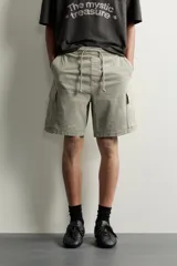 Bermuda cargo color negro de corte relaxed fit, confeccionada en tejido de algodón con elasticidad y efecto lavado. Cuenta con cintura elástica ajustable con cordón, bolsillos frontales y bolsillos laterales tipo cargo con solapa en las perneras.
