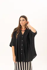 Camisa negra de corte oversize con diseño de encaje calado, escote en V, cierre frontal con botones y mangas tres cuartos. Presenta un diseño asimétrico con la parte posterior ligeramente más larga.