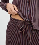 Pijama de dos piezas compuesto por una camisa de manga larga con cuello clásico, cierre frontal de botones y detalle de ribete, y un pantalón largo de corte recto. Ambas prendas presentan un diseño de rayas verticales finas en tono oscuro.