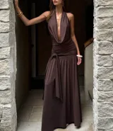 Vestido largo color marrón chocolate, con escote halter profundo y drapeado en el busto. La cintura es ajustada con frunces y un lazo anudado, y la falda es fluida con una abertura lateral.