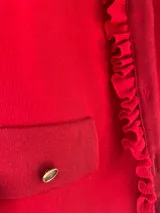 Cardigan de lana merino color rojo, con cuello redondo, mangas largas y cierre frontal de botones dorados. Presenta detalles de volados a lo largo de la botonera y bolsillos frontales simulados con solapa y botón.