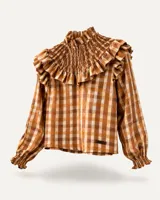 Blusa de manga larga con estampado de cuadros en tonos marrones y beige. Presenta cuello alto con detalle de fruncido elástico (smock) y volados decorativos en la parte superior del pecho y hombros. Las mangas terminan en puños elásticos con volados.