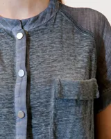 Remera blanca de lino con cuello mao, media cartera con broches metálicos y manga japonesa. Presenta un bolsillo frontal tipo plaqué, recortes laterales en sisa con detalles de encaje y ruedo redondeado con corte escalonado.