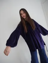 Buzo tipo canguro oversize de tela plush, color azul claro. Presenta capucha, cierre frontal parcial con cordón turquesa y bolsillos canguro frontales. Las mangas son largas y abullonadas con puños ajustados.