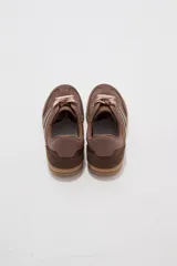 Zapatillas urbanas de corte bajo en tonos beige y tostado, combinando paneles de cuerina y gamuza sintética. Cuentan con cordones anchos tipo cinta de raso en color crudo, pespuntes decorativos y suela de goma al tono.