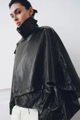 Chaqueta de efecto piel en color negro con cuello subido y bufanda desmontable integrada mediante botones. Presenta manga larga con trabillas ajustables en los puños, cierre frontal con cremallera y bolsillos laterales ocultos en las costuras. El diseño incluye un bajo con elástico en la espalda y forro interior.