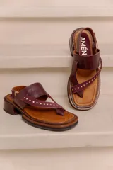 Sandalia de cuero bordó con diseño tipo thong, tiras anchas cruzadas con tachas metálicas doradas, tira con hebilla ajustable en el talón y taco bajo cuadrado con acabado artesanal.