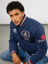 Campera bomber azul marino estilo retro, con cierre frontal de botones a presión, cuello, puños y ruedo con tejido acanalado tricolor. Presenta el logo de Umbro y el escudo del Club Nacional de Football bordados en el pecho, junto con el año 1899 y parches decorativos en las mangas.