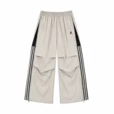 Pantalón deportivo baggy beige impermeable con cierre completo en los laterales, bandas negras con tres líneas blancas a los costados y cintura elástica ajustable.