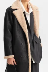 Abrigo largo de color negro con interior de corderito en contraste beige. Presenta un diseño de cierre cruzado con botones, solapas anchas y bolsillos laterales.