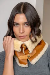 Cuello de abrigo confeccionado en cuero ovino color suela, con interior y bordes de piel sintética en tono crema. Presenta cierre frontal con botones a presión metálicos y tiras de ajuste.