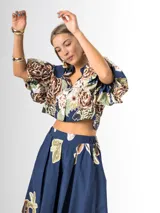 Conjunto de dos piezas confeccionado en viscosa con estampado floral en tonos marrones y verdes sobre base blanca. Incluye una blusa tipo camisa corta con cuello solapa, cierre frontal de botones y mangas abullonadas con diseño globo, y un pantalón tipo culotte de tiro alto, amplio y fluido con pinzas en la cintura.