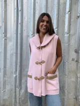 Chaleco de lana merino color rosado, con cuello alto tejido, cierre frontal tipo duffle con botones de madera y detalles en cuero sintético en los bolsillos y cierres.