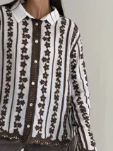 Camisa blanca de manga larga con delicados bordados florales verticales y detalles de encaje. Presenta cuello clásico, cierre frontal con botones y terminaciones festoneadas en los puños y el ruedo.