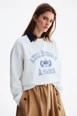 Buzo oversize color blanco de cuello redondo y mangas largas, con interior cepillado suave. Presenta un estampado frontal en tono azul con texto de estilo universitario y diseño decorativo. Cuenta con ribetes acanalados en el cuello, los puños y el bajo.