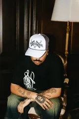 Gorra trucker gris con paneles frontales blancos y paneles traseros de rejilla negra. Presenta un logo bordado en el panel frontal y una cuerda decorativa en la base de la corona.