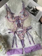 Body de lencería de tul transparente en tono lila con detalles de encaje bordado floral en tonos violeta y celeste, aberturas estratégicas y portaligas integrados con tiras ajustables.