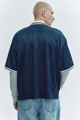 Camiseta de fútbol de manga corta en malla estampada, con diseño a rayas verticales en tonos azul y verde oscuro. Cuenta con cuello polo blanco y escote en V con ribete negro, además de estampados gráficos frontales con estrellas y la inscripción "Run Wild". Presenta un corte holgado.