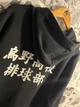 Campera negra con capucha y cierre frontal, con estampado en la espalda de texto en japonés en color blanco. Diseño inspirado en el anime Haikyu!!