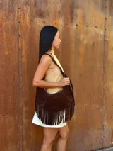 Cartera de hombro tipo hobo confeccionada en cuero gamuzado color chocolate, con detalle de flecos largos en la parte inferior y correa ajustable con hebilla metálica.