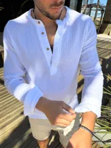 Camisa de muselina blanca con cuello mao y cierre frontal parcial de botones. Presenta mangas largas con detalle de trabilla para arremangar y textura suave.
