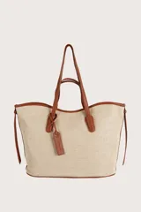 Bolsa tipo tote de textura tejida en color natural con detalles y correas en cuero sintético color suela. Presenta un diseño amplio con doble asa de hombro y un dije colgante decorativo.