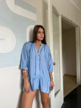 Vestido corto azul claro de manga larga, con cuello mao y cierre de botones frontal. Presenta detalles de encaje o bordado en las costuras verticales del frente y en la zona de los hombros. La manga se ajusta con lazo y el ruedo tiene un volante fruncido.